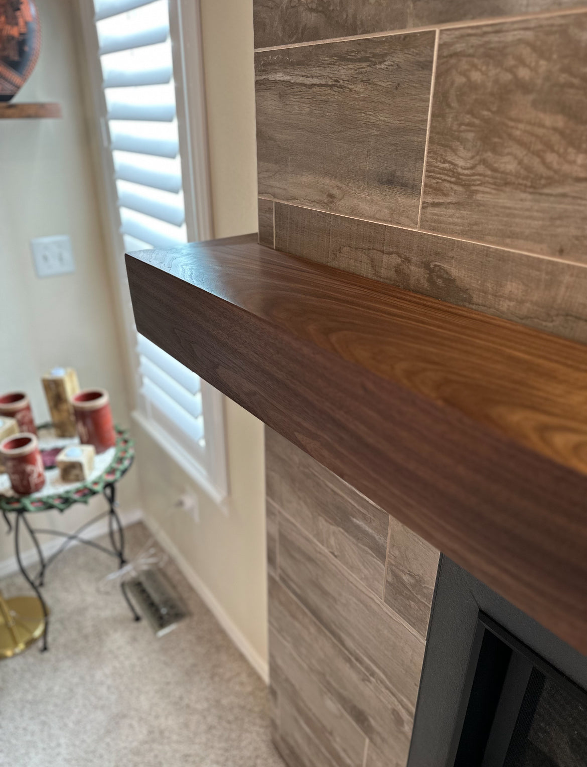 Wooden mantel against a tiled wall with a small table and decor items in the background.
